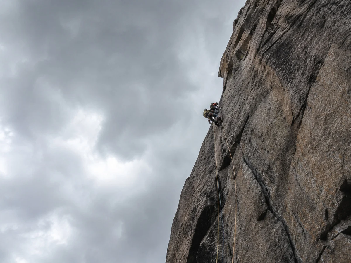 A climber partway up a grey multi-pitch wall under heavy cloud